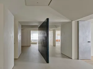 Modern minimalist hallway featuring light micro-cement walls and floors, a tall dark metal pivot door, and views into adjacent living and dining areas.