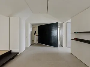 Minimalist Japanese-style entryway featuring a large black pivot door, micro-cement walls and floors, built-in storage, and floating dark shelves with a woman in white standing in the background.
