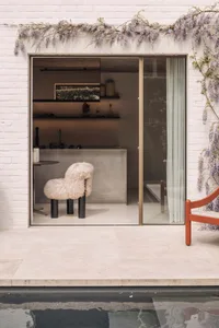 Modern minimalist outdoor kitchen with concrete island, black cylindrical stool covered in cream Mongolian fur, open shelving, and blooming wisteria vine framing the white brick exterior.
