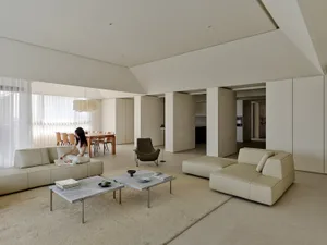 Spacious modern living room with beige leather sofas, micro-cement walls, concrete coffee tables, and a woman reading on the sofa in natural daylight.