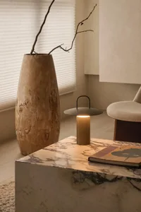 Close-up of a luxurious Calacatta marble coffee table featuring a lit portable lantern, a large textured ceramic vase with dried branches, and a hardcover book, in a minimalist neutral-toned living room.