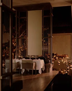 Elegant fine-dining restaurant interior featuring dark wooden lattice room dividers, white tablecloth dining tables, and golden leaf wall art under warm lighting.