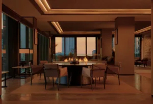Modern luxury dining room featuring a large round walnut dining table with six upholstered chairs, warm cove lighting, and floor-to-ceiling windows showing a city skyline at sunset with a fireplace in the background.