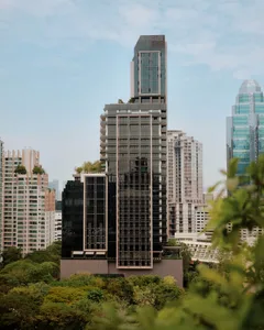 高聳的現代住宅摩天大樓，深色玻璃帷幕與垂直線條，在藍天下被茂密綠樹與其他城市建築環繞。