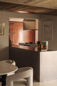 Modern minimalist kitchen featuring a concrete island, warm walnut wood cabinetry, wood-paneled ceiling, and sculptural boucle dining chairs.