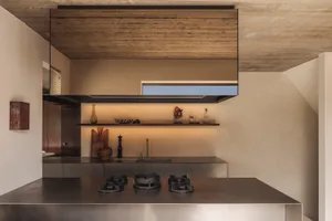 Modern minimalist kitchen featuring raw concrete surfaces, stainless steel island with gas burners, floating wooden ceiling element, and backlit open shelving displaying decorative objects.