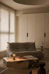 Modern minimalist living room featuring a grey Togo sofa, a large Calacatta Viola marble coffee table with a lit candle and book, wooden stool, and curved ceiling detail.