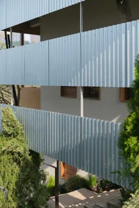 Modern residential building exterior featuring light blue corrugated metal cladding, wooden louvered windows, and surrounding evergreen trees and shrubs.