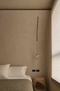 Minimalist bedroom corner featuring a textured travertine wall, white bedding, a hanging crystal pendant light, and a solid wood stump stool beside a window with sheer blinds.