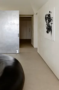 Modern minimalist hallway featuring seamless micro-cement walls, concrete floor, a large black sculptural object in the foreground, and a black and white abstract engine artwork hanging on the right wall.