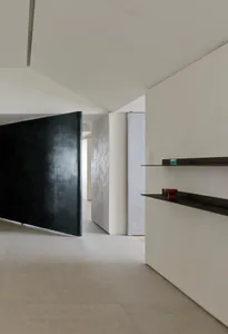 Modern minimalist hallway featuring a large black pivoting door, light micro-cement walls, floating black shelves with decorative objects, and seamless concrete flooring.