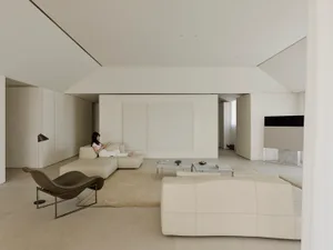 Spacious minimalist living room with micro-cement walls, large off-white sectional sofa, brown leather lounge chair, and a woman reading on a modular sofa.