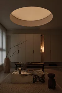 Minimalist living room featuring a large marble coffee table, low wooden bench, textured beige walls, and a dramatic circular ceiling light fixture.