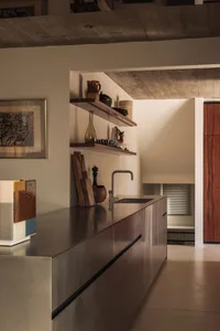 Modern minimalist kitchen featuring a long stainless steel countertop and cabinetry, open wooden shelves displaying ceramics, concrete ceiling, and a framed abstract artwork on the wall.