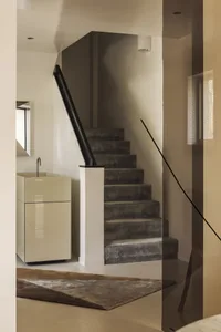 Modern minimalist entryway featuring a floating dark velvet carpeted staircase with a bold black handrail, paired with a glossy cream vanity and geometric rug.