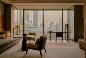 Modern luxury living room with curved walnut table, taupe leather armchairs, built-in sofa, and dramatic floor-to-ceiling windows overlooking the Hong Kong skyline at dusk.