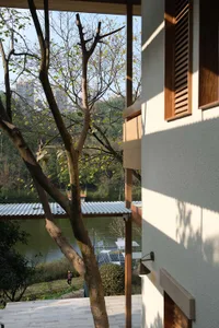 View from a modern house terrace featuring wooden window frames and shutters, overlooking a lake with trees and outdoor seating area below.