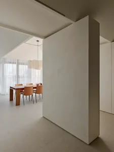 Modern minimalist dining room featuring a large micro-cement partition wall, wooden dining table, tan leather chairs, and a sculptural pendant light under a high sloped ceiling.
