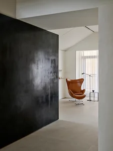Modern minimalist living room featuring a large black micro-cement accent wall, a brown leather Egg Chair with ottoman, and natural light from a tall window with sheer curtains.