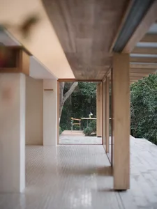 View from inside a minimalist covered terrace with wooden beams and concrete tile floor looking out to a lush green garden with wooden deck and furniture.