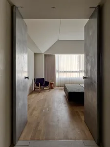Minimalist bedroom interior with raw concrete walls and doors, light oak flooring, sloped ceiling, low platform bed with dark linen bedding, and a blue armchair by the window.
