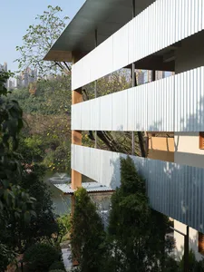 Modern hillside residential building featuring white corrugated metal cladding, warm timber columns, and large overhangs overlooking a garden pond with fountain surrounded by lush greenery.