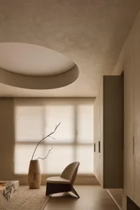 Minimalist living room interior with beige micro-cement walls, curved ceiling feature, beige upholstered reading chair, wooden vase with dried branch, marble coffee table, and textured area rug.