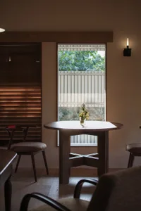 Modern minimalist dining area featuring a solid walnut round table, matching wooden chairs with upholstered seats, warm wood slat wall, and a large window with garden view.