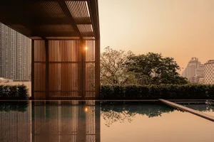 Modern rooftop infinity pool reflecting a dramatic orange sunset, framed by vertical wooden slat screens and a city skyline in the background.