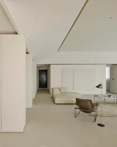 Minimalist living room featuring off-white micro-cement floors and walls, a low-profile cream leather sectional sofa, a dark olive sculptural lounge chair with chrome frame, and a slim floor lamp.
