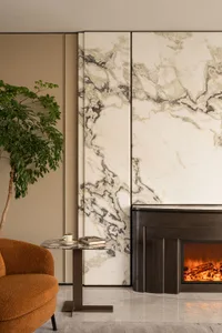 Modern living room featuring a large Calacatta marble feature wall, dark metal fireplace with burning fire, burnt orange bouclé armchair, marble side table, and a tall potted tree.