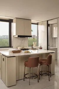 Modern minimalist kitchen featuring a large beige island with white Calacatta marble countertop, two brown leather bar stools, handleless cabinetry, and large windows overlooking greenery.