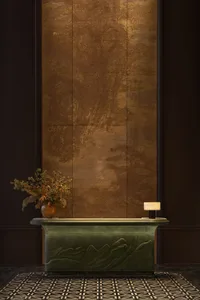 Luxury hotel reception desk in verdigris green with mountain relief carving, placed against a textured gold leaf wall with dried floral arrangement and marble lamp.
