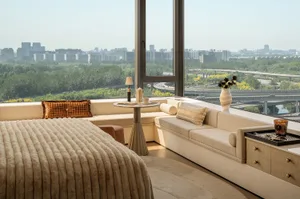 Modern high-rise bedroom featuring a large boucle fabric bed, built-in window seat with orange accent pillows, round side table, and floor-to-ceiling windows overlooking a vast cityscape with green areas and highways.