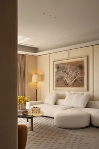 Modern luxury living room with a creamy white boucle L-shaped sectional sofa, round ottoman, marble coffee table, floor lamp, and large framed abstract tree artwork on a textured beige wall.