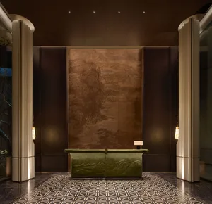 Luxurious hotel reception desk featuring a carved green marble console table against a large bronze panel with traditional Chinese landscape artwork, flanked by tall beige columns and geometric patterned marble floor.