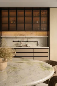 Modern dining room with round Calacatta green marble table, beige curved dining chairs, and a luxurious marble wet bar with amber textured glass upper cabinets and under-cabinet lighting.
