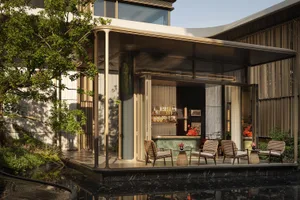 Modern luxury courtyard bar with brass-framed glass pavilion, green marble counter, rattan lounge chairs, and a dark marble reflecting pool surrounded by trees.