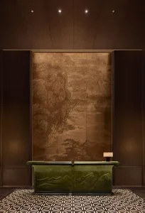 Luxurious hotel or corporate reception desk featuring a carved malachite-green front with mountain relief, set against a large bronze-toned Chinese landscape mural, under a geometric coffered ceiling with patterned marble floor.