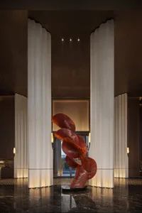 Luxurious hotel lobby featuring a large abstract red sculpture with gold detailing, flanked by tall white fluted marble columns on a polished dark marble floor.