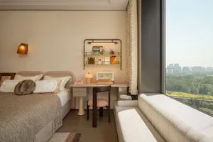 Modern serene bedroom featuring a pink boucle headboard, textured beige bedding, built-in vanity desk with floating shelves, and a large window with city view.