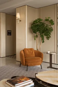 Modern living room corner featuring a burnt orange boucle armchair, tall potted tree, marble side table, and textured beige walls with wall sconce.