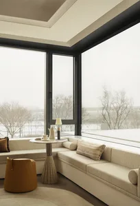 Modern minimalist living room corner featuring a built-in boucle fabric window seat banquette, round fluted side table, mustard yellow leather ottoman, and large black-framed windows with winter view