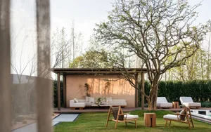 Modern outdoor living space featuring wooden lounge chairs, white sectional sofa, peach-colored wall, and a large deciduous tree on a manicured lawn.