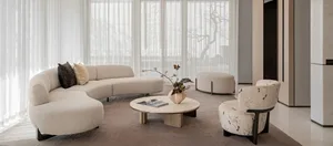 Modern minimalist living room featuring a large cream boucle curved sectional sofa, matching floral accent armchair, round light-wood coffee table, and sheer white curtains allowing abundant natural light.