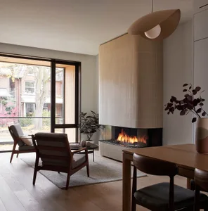 Modern minimalist living room featuring a tall ribbed-tile fireplace with burning fire, walnut wood armchairs, wooden dining table, and large glass doors opening to a garden.