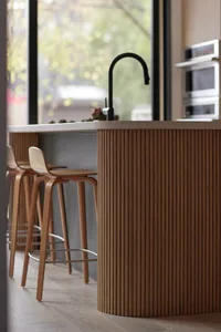 Modern kitchen island featuring a curved fluted oak veneer base, white countertop, black gooseneck faucet, and three light wood bentwood bar stools in front of large windows.