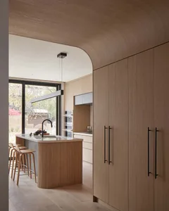 Modern minimalist kitchen with light oak cabinetry, a curved wooden ceiling, fluted oak kitchen island with white countertop, and wooden bar stools.