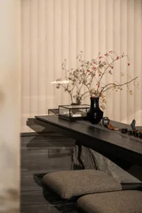 Minimalist wabi-sabi style interior featuring a dark wooden console table with natural stone base, adorned with a black ceramic vase holding red berry branches, incense burner, and two floor cushions on polished dark marble floor.