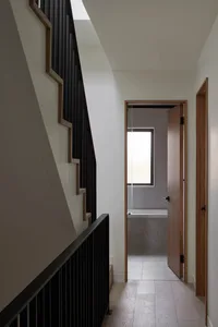 Modern minimalist hallway with black wooden staircase on the left and an open doorway leading to a bathroom with natural light at the end.